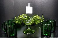 Full frontal view of the candy bowl surrounded by five matching green glass mugs in a single line on either side, showing fluted edges and pedestal base with smooth glossy glass.