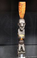 Front view of the statue showing the male figure kneeling with hands clasped in prayer, detailed facial features, and a tall elongated orange headdress with yellow tribal patterns.