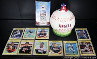 Photo showing the ceramic baseball trinket box with red stitching, the 'ANGELS' logo, green base, the 2002 World Champion pin in packaging, and 10 Anaheim Angels 1985 Fleer trading cards laid out in two rows in front.