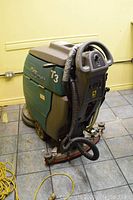 Perspective view of floor scrubber on tiled floor showing handle, scrub deck, hose and wheels