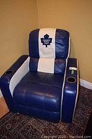 Full view of blue and white leatherette recliner with Toronto Maple Leafs logo headrest, two black cup holders, placed on patterned rug in front of tan wall