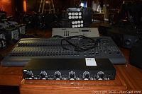 Foreground speaker selector unit with eight volume knobs; midground grey rackmount box and white lighting console; background cube power distro and black gear.