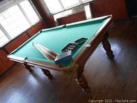 Three-quarter view of full pool table showing playing surface, cues, rack on felt and room setting