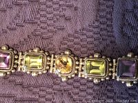 Close-up photo showing five linked square and rectangular shaped colored gemstones in purple, amber, and yellow tones set in sterling silver with decorative beaded design.