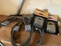 Two braided steel security cables with attached silver locks, and two Master Lock padlocks in original packaging displayed on wooden surface.