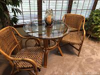Two rattan chairs shown with a glass table in a room with carpet and plants.