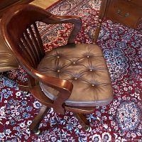 Front angle of cherry wood swivel office chair with tufted leather cushion and armrests on a patterned rug.