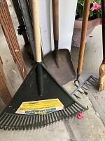 Photo showing a green plastic leaf rake, metal square shovel, metal rake, and small broom with wooden handles, placed on a concrete floor near a potted plant.