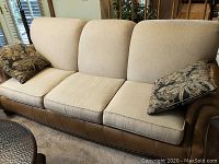 Front view of three-seat sofa with beige fabric cushions, brown leather base, and two paisley throw pillows