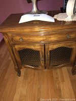 Photo of one wooden nightstand showing wood grain, drawer with round metal pull, and double cabinet doors with black metal mesh inserts.