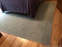Photo showing large sage green rug placed beside bed on wood floor with visible intricate floral and vine border design.