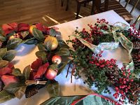 Two Christmas wreaths laid out on a table, showing the variety of decorations including large red flowers, green leaves, pinecones, faux fruits, and a red and gold ribbon.
