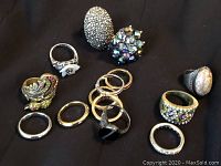 Overview of assorted costume rings including rhinestones, flower detail, and plain bands on black background.