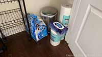 Wide view of trash can and surrounding paper products on the floor showing papers towels and bathroom tissue rolls