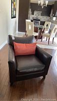 Front angled view of the dark brown faux leather armchair with orange lumbar pillow set in a home environment showing wood flooring and dining table area in background.