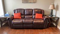 Full front view of the brown leather three-seater recliner sofa with two orange pillows on it.