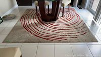 Top-down view of the square rug with red swirl pattern under a dining table, showing the full rug and signs of wear and dirt.