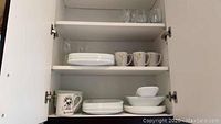 White Corelle plates, bowls, and mugs in cabinet shelf showing multiple pieces, some decorative print mugs.