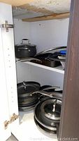 Cupboard interior showing stacked pots and pans, including large black pot with lid, frying pans and grill pan.