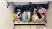 View of cleaning supplies under kitchen sink showing various bottles, spray cleaners, wipes container, paper towels, and cleaning brushes.