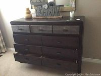 Front view of dark wood dresser with nine drawers, three rows of three drawers each. Top row drawers have fabric-front panels with dark handles. Surface holds decorative items not included.