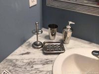 Photo showing four polished stainless steel bathroom accessories on marble countertop: toothbrush holder, cup, soap dish, pump bottle