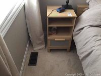 Night stand next to bed showing one drawer and two open shelves in light wood with plastic panel on drawer front