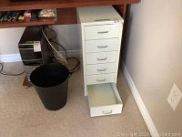 White metal drawer cabinet with six drawers, one drawer open, front view showing metal handles. Cabinet measures 11x16x26 inches. Black resin waste basket beside cabinet on floor.