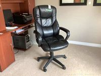 Front view of black faux leather office chair with armrests and wheel base, positioned on carpeted floor near wooden desk.