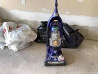 Full front view of purple Bissell upright vacuum cleaner in garage area with trash bags behind it.
