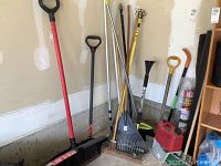 Photo showing snow shovel, brooms, rakes, ice chopper, and other long handled garden tools stored upright against a wall on a garage floor.