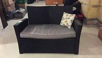 Front view of black synthetic wicker loveseat with gray cushion and patterned throw pillow