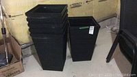 Six black square resin planters stacked and standing side by side on a concrete floor in a basement or storage area.