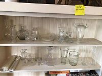 Shelf with assorted clear glass vases and candleholders of various shapes and sizes, with small bowls and glasses.