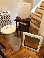 Vintage wood side table with white fabric shade table lamp on top, white metal can with lid and a translucent wastebasket in front, and white metal framed mirror leaning against wall.