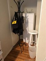 White metal shoe storage bin, white metal stool, black wrought iron coat rack, and plastic wastebin shown positioned in corner of room on wood floor.