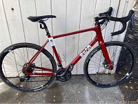 Full left-side profile of red Bear Bigrock gravel bike leaning against white wall.