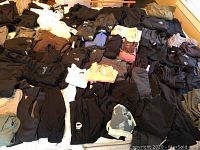 Image shows a large assortment of folded women's activewear garments including tops, leggings, and sports bras, predominantly in black and gray tones with some olive and peach items.