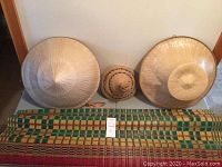 Full view of the woven grass mat laid horizontally with three straw hats placed above it against a wall, showing the size and condition of all items