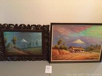 Photo of two paintings side by side on a wall: left is smaller canvas painting with carved wooden frame showing Cotopaxi volcano landscape; right is larger Masonite painting with village and volcano under colorful sky, both framed in wood.