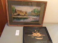 Photo of two framed art pieces side by side. Larger is watercolor with naturalistic rural scene, smaller is rice straw on Thai silk depicting boat figures.