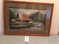 Full view of the framed watercolor painting showing the rural scene with thatched huts, water, and a boat.