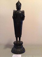 Front view of black metal standing Buddha statue on pedestal, both hands raised in teaching mudra.