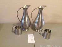 Full view of two pewter pitchers and two small cups arranged on a surface to show the entire set