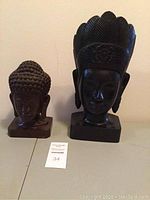 Two carved wooden heads side by side, one lighter Buddha head and one darker head with headdress on flat surface.