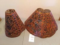 Full view of two tightly curled pierced leather lampshades with detailed etched figures and patterns.