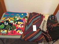 Image showing colorful textile items including two 3-D fabric wall hangings, one folded red and black woven skirt, and a black embroidered top with pom-poms and a cream-colored woven top.