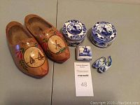 Photo showing the full lot with two round blue and white porcelain trinket boxes, one small square porcelain box with windmill design, a pair of tiny blue and white porcelain shoes, and a pair of large wooden Dutch shoes decorated with painted scenes and 'Holland' written on them.