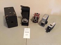 Full view of all five items including box camera, Rolfix Jr folding camera, Rondine Ferrania camera, and two flash attachments