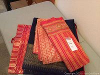 Three folded pieces of Thai fabric including midnight blue silk with gold thread, pale red and gold silk, and pink and green cotton fabric sewn into a tube shape, laid on a surface